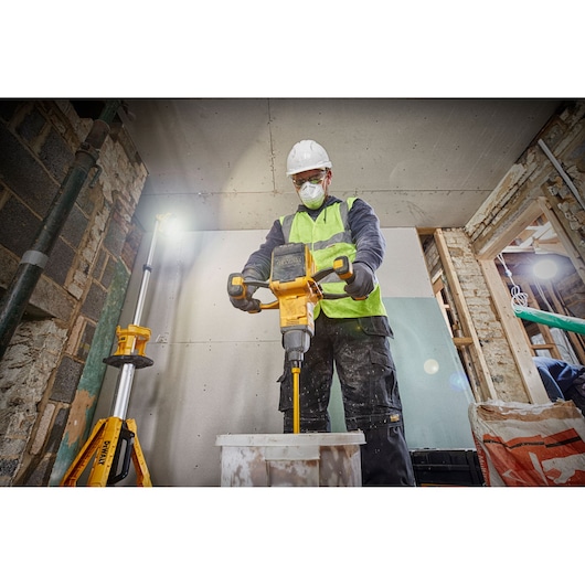 Worker mixes materials with a DEWALT tool indoors, wearing safety gear and a high-visibility vest.