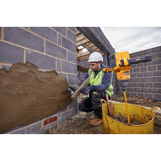 A construction worker wearing safety gear applies mortar to a brick wall using a trowel. Next to the worker, a yellow DEWALT DCD240N mixer is attached to a mixing paddle in a large yellow bucket with mortar. The scene is set at a construction site with unfinished brick walls.