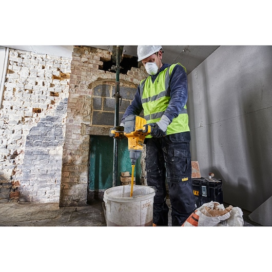 A person wearing a safety vest and gloves is using a DEWALT DCD240X2 cordless mixer to stir contents in a large bucket at a construction site with brick and drywall walls.