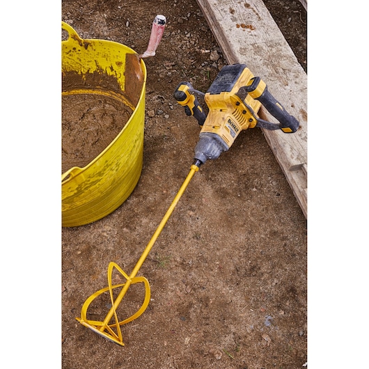 A DEWALT cordless mixer with a long yellow mixing paddle is lying on a construction site floor next to a yellow bucket containing mixed material and a trowel.