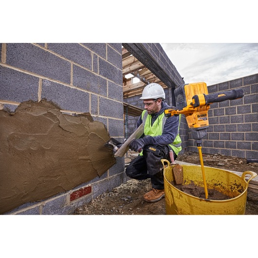 A construction worker wearing a safety helmet and a high-visibility vest applies plaster to a brick wall at a building site. Next to the worker is a yellow DEWALT mixing drill (model DCD240X2) placed in a large yellow container filled with plaster mix.