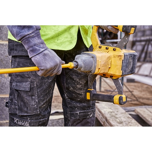 A person wearing work gloves and protective clothing is holding a DEWALT DCD240X2 power mixer, using it to mix construction material on a job site.