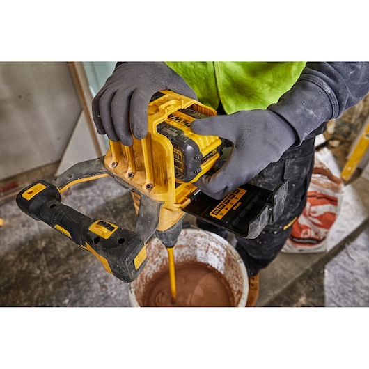 A person using a DEWALT cordless mixer to mix material in a large bucket on a construction site. The person is wearing work gloves and a high-visibility jacket.