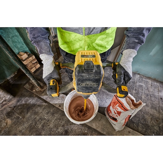 A person wearing work gloves is using a DEWALT cordless mixer to mix material in a white bucket on a construction site. A bag of material is open next to the bucket.