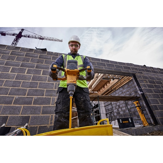 A construction worker wearing a high-visibility vest and a safety helmet is using a yellow DEWALT power mixer on a construction site with partially built brick walls. A crane is visible in the background and DEWALT equipment can be seen nearby.