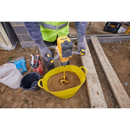 A construction worker is using a yellow DEWALT mixing drill to mix concrete or mortar in a large yellow bucket outdoors. The worker is wearing gloves and a high-visibility vest, with other buckets and bags of materials nearby on the ground.
