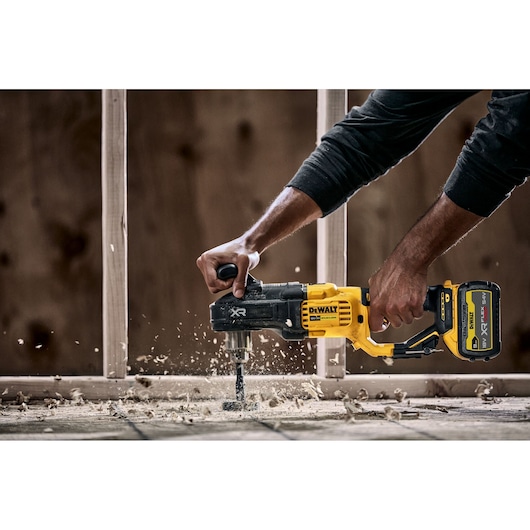 A person using a DEWALT DCD444N cordless power drill with a side handle to drill through wood, creating wood shavings and dust. Construction framing is visible in the background.