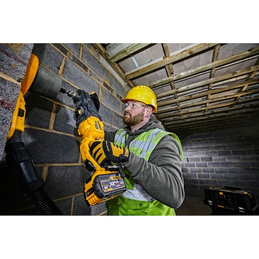 A person wearing a yellow hard hat, safety gloves, and a high-visibility vest is using a DEWALT cordless drill (DCD470N) to bore a hole into a concrete block wall at a construction site. The person's face is blurred.
