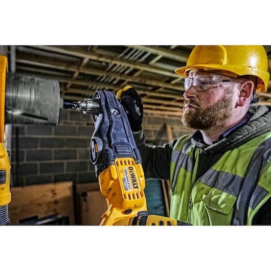 A person wearing a yellow hard hat and safety vest is using a DEWALT DCD470N power drill with an attachment to drill into a surface at a construction site. The person’s face is blurred for privacy.