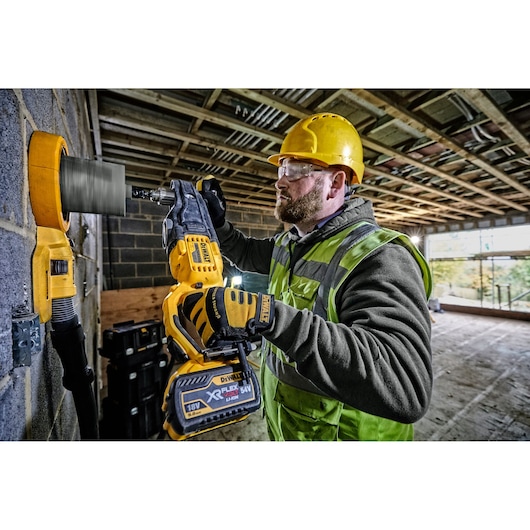 A construction worker wearing a yellow hard hat and high-visibility vest uses a DEWALT DCD470N cordless drill with a hole saw attachment to drill into a concrete wall at a building site.