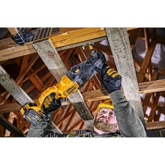 A person using a DEWALT DCD470N cordless drill to drill a hole in a wooden beam as part of construction work. The worker is wearing protective gloves and a safety helmet.