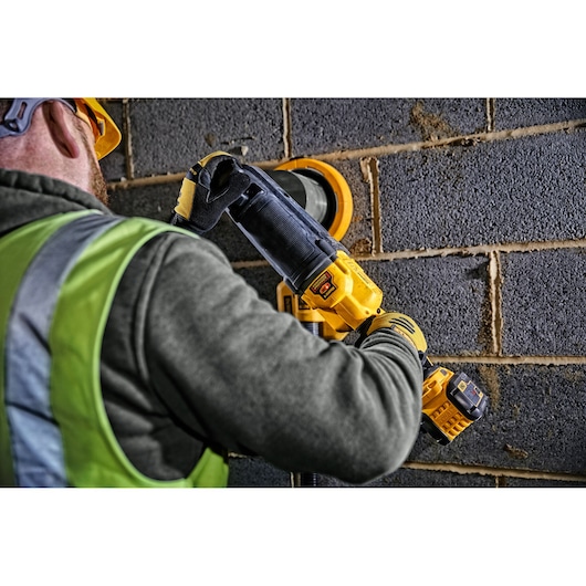 A person wearing a safety vest and helmet is using a DEWALT DCD470N cordless drill against a brick wall in a construction setting.