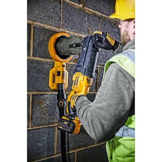 A construction worker wearing safety gear is using a DEWALT cordless drill with a hole saw attachment to drill into a brick wall. The tool has a vacuum system attached to collect dust during the operation.