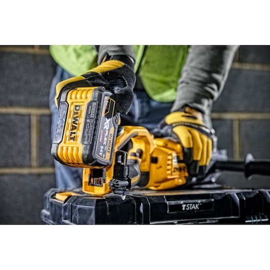 A person wearing work gloves is inserting a DEWALT XR FLEXVOLT 54V battery into a yellow DEWALT power tool, positioned on a black TSTAK storage box, with a brick wall in the background.