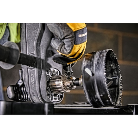 Close-up of a person wearing yellow and black work gloves using a DEWALT power tool equipped with a large hole saw drill bit, preparing for heavy-duty drilling on a work surface.
