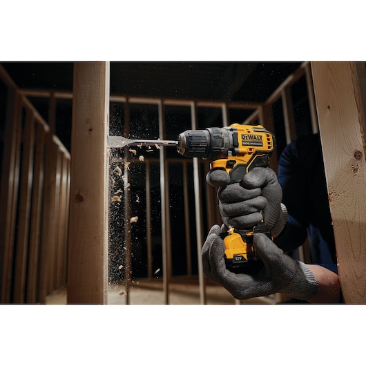 A person wearing protective gloves uses a yellow DEWALT cordless drill to drill into a wooden beam inside a framed structure. Wood chips are visible flying from the drilling process.