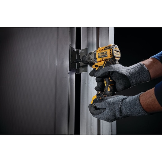 A person wearing gray gloves uses a yellow and black DEWALT cordless drill to secure a hinge on a metal door frame.