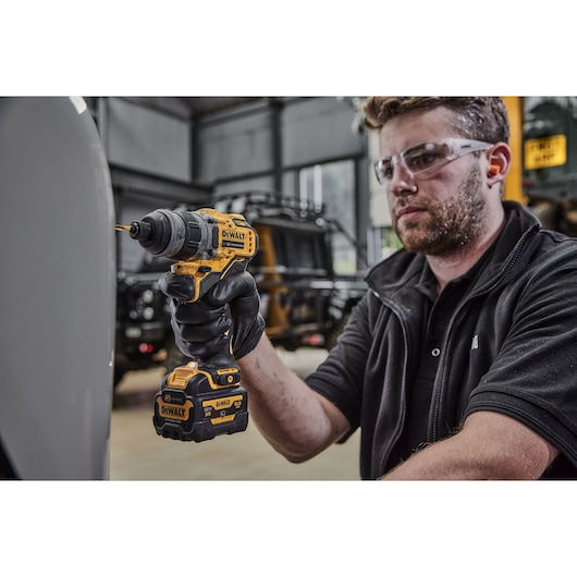 A person wearing black gloves is using a yellow and black DEWALT cordless drill with a battery attached indoors. The face of the person is blurred for privacy.