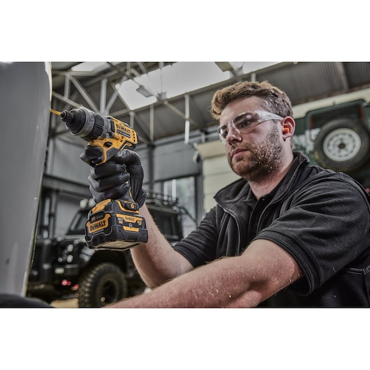 A person with a blurred face is using a DEWALT DCD703NT cordless power drill in a workshop or garage setting. The drill is yellow and black, and the person is wearing black gloves.