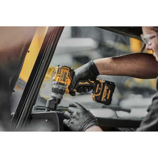 A person wearing black gloves uses a yellow and black DEWALT cordless drill with a brushless motor to work inside a vehicle near a window.