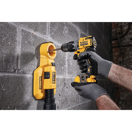 A person wearing gray gloves is using a yellow DEWALT cordless drill to drill into a concrete wall, with a yellow dust extractor attached to collect debris.