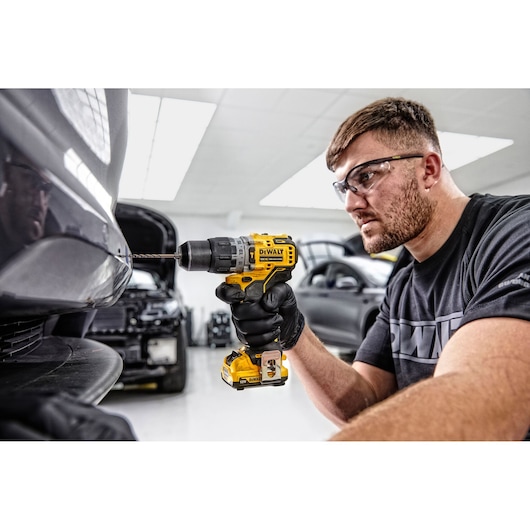 A person wearing black gloves uses a yellow DEWALT DCD706D2 cordless drill to drill into the bumper of a car in an automotive workshop. The person's face is blurred and several cars are visible in the background.