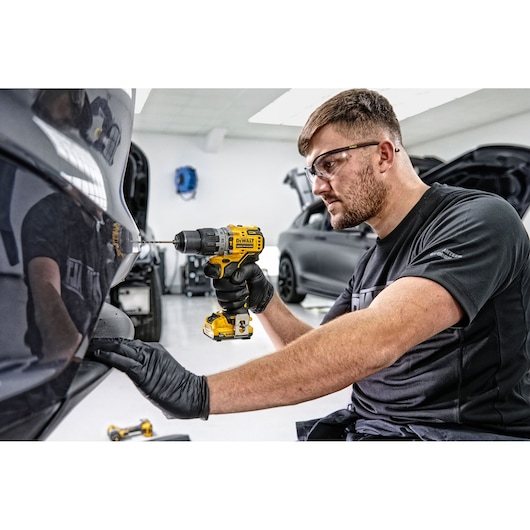 A person wearing black gloves uses a DEWALT cordless drill on the rear bumper of a car in a workshop setting. The person's face is blurred for privacy.