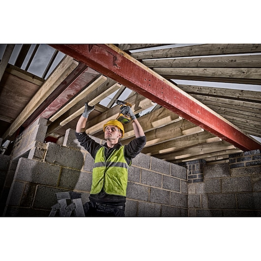 A construction worker wearing a yellow hard hat and high-visibility vest uses a DEWALT cordless drill to secure wooden beams to a red steel support inside a building under construction. The face of the worker is blurred.