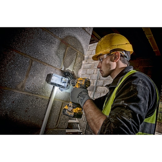 A person wearing a yellow hard hat and safety vest is using a DEWALT DCD796P2 cordless drill to work on a metal electrical box mounted on a brick wall. The person's face is blurred for privacy.