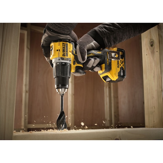Gloved hands using a DEWALT cordless drill to bore into wood, producing shavings as it drills.