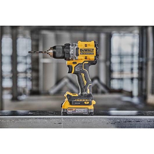 A DEWALT 18V XR Brushless cordless drill driver with a drill bit attached, standing upright on a metal surface in an industrial workspace. The tool features black and yellow colors and POWERSTACK battery.