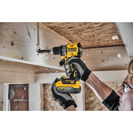 Person drills wood beam with DEWALT cordless drill, wearing gloves and safety glasses in a workshop.