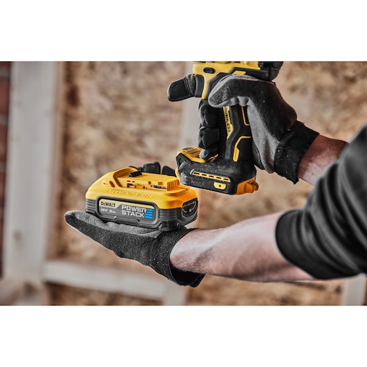 Gloved hands hold a DEWALT drill and 18V Power Stack battery in a workshop environment.