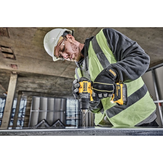 A person wearing a safety vest, gloves, and a hard hat is using a DEWALT DCD800NT cordless drill to drill into a metal surface at a construction site.