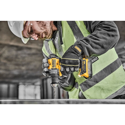 A person wearing safety gear, including a helmet, goggles, gloves, and a high-visibility vest, is using a yellow and black DEWALT cordless power drill at a construction site.