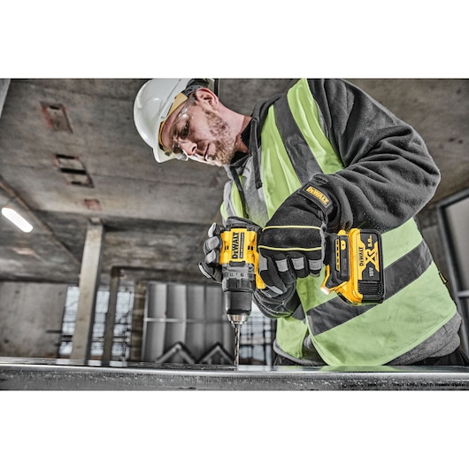 A person wearing safety gear, including gloves, a helmet, and a hi-vis vest, is using a DEWALT DCD800P2T cordless drill to work on a metal surface in an industrial environment.