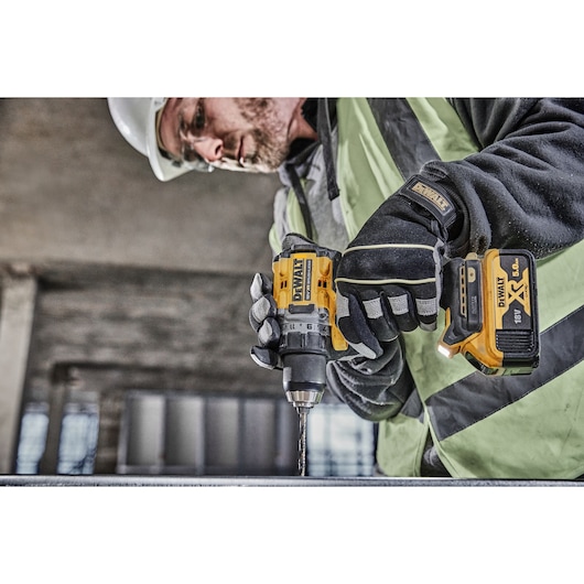 A person wearing safety gear operates a DEWALT DCD800P2T cordless drill with an 18V XR battery, drilling into a metal surface at a construction site.