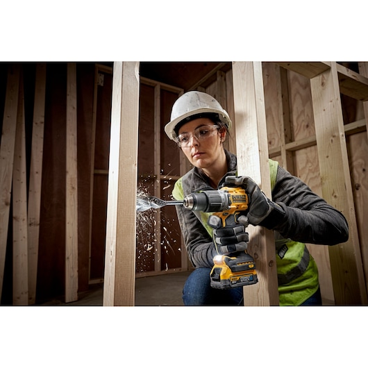 Person drills wood with a DEWALT DCD805E2T tool at a construction site. Safety gear and gloves are worn.