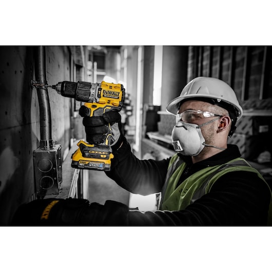 Worker uses DEWALT DCD805E2T cordless drill on a metal pipe. Wearing safety gear in an industrial setting.