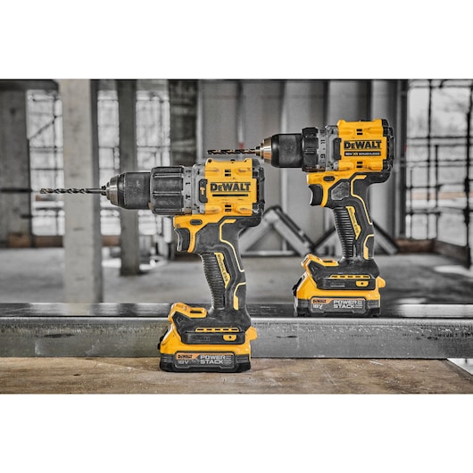 Two DEWALT cordless power drills with yellow and black design, standing on a metal surface in an unfinished indoor construction environment. Both drills feature 18V POWERSTACK batteries.