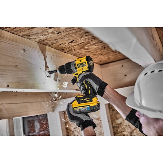 Person uses DEWALT cordless drill to make a hole in wooden beam, wearing gloves and hard hat.