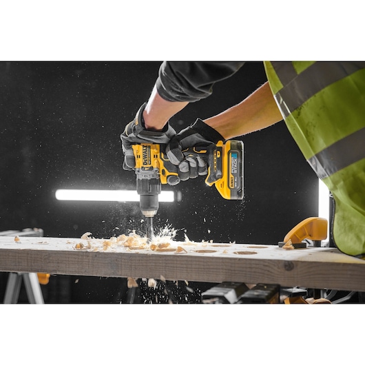 Gloved hands use a yellow DEWALT drill to work on a wooden plank, creating wood shavings and sparks.
