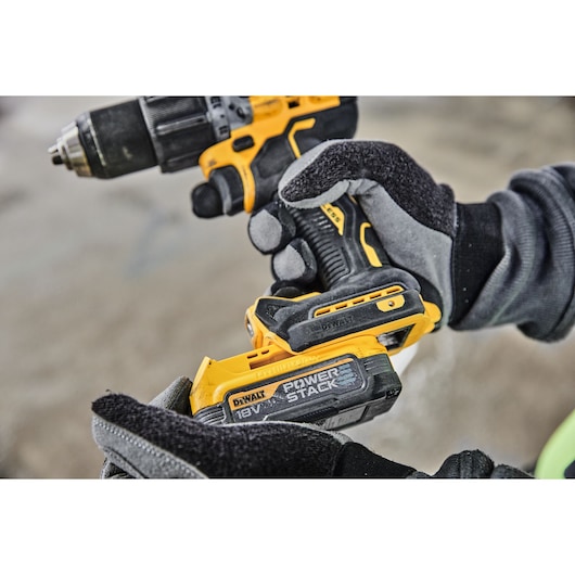Close-up of gloved hands holding a yellow and black DEWALT cordless drill, with visible 18V POWERSTACK battery being attached.