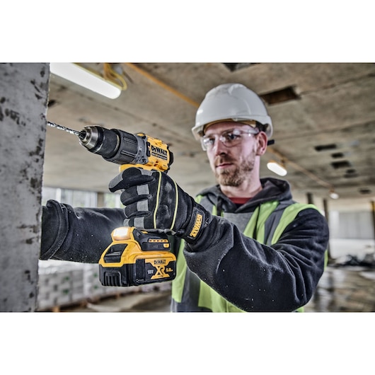 A person wearing gloves, a safety vest, and a hard hat is using a DEWALT DCD805P2T cordless drill to drill into a concrete surface at a construction site. The face is blurred for privacy.