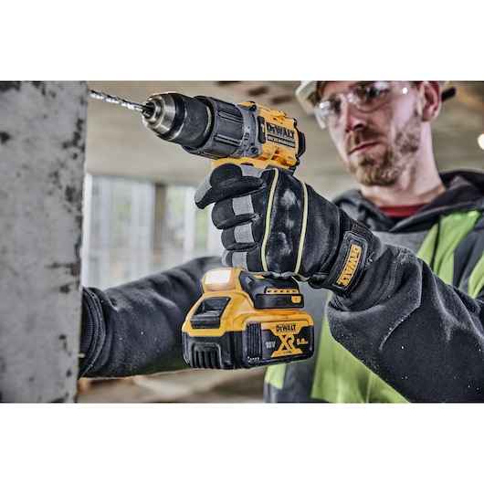 A person wearing DEWALT-branded gloves is using a DEWALT 18V XR cordless drill driver (model DCD805P2T) to drill into a concrete surface in an indoor construction setting. The face of the person is blurred.