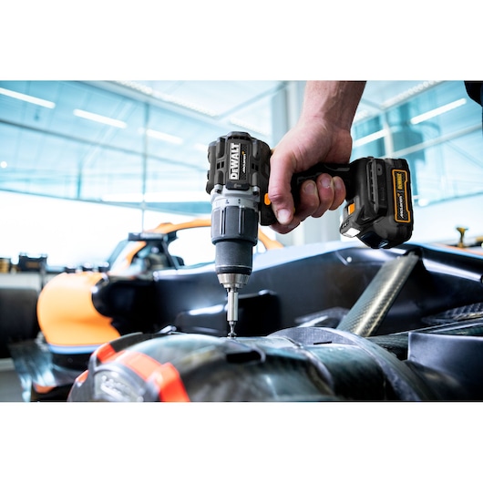A close-up of a DEWALT cordless drill being used by a person to work on a part of a vehicle, with an orange car visible in the background in a bright, modern workshop.
