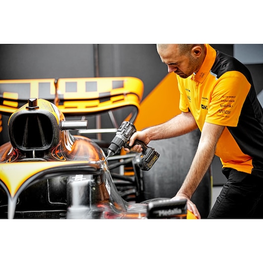 A person in an orange and black shirt is using a DEWALT cordless drill on a Formula 1 race car. The focus is on the DCD85ME2GT power tool and the car's aerodynamic components.