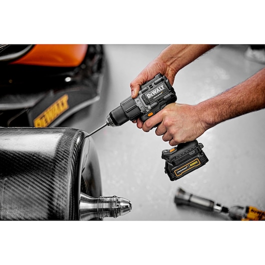 A person uses a DEWALT cordless power drill, model DCD85ME2GT, to drill into a sleek, carbon fiber surface. The drill has a black and yellow design and is battery powered.