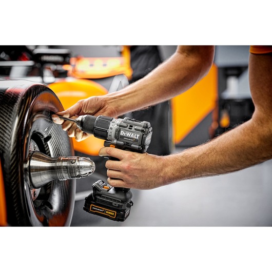 A person uses a DEWALT cordless drill (model DCD85ME2GT) to work on the wheel of a race car in a workshop setting.