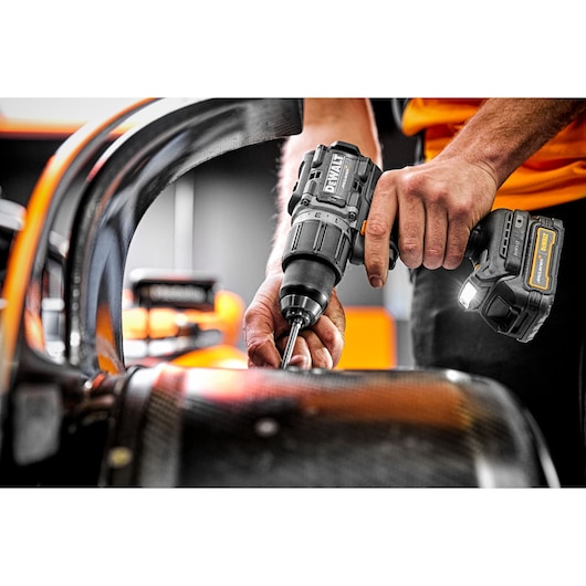 A close-up of a person using a DEWALT cordless power drill on a carbon fiber surface, with a sports car blurred in the background.
